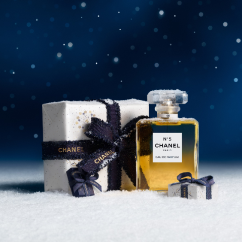 Ein Flakon des Parfums Chanel No. 5 steht auf Schnee neben zwei verpackten Geschenken mit marineblauen Bändern vor einem dunkelblauen, verschneiten, sternförmigen Hintergrund.