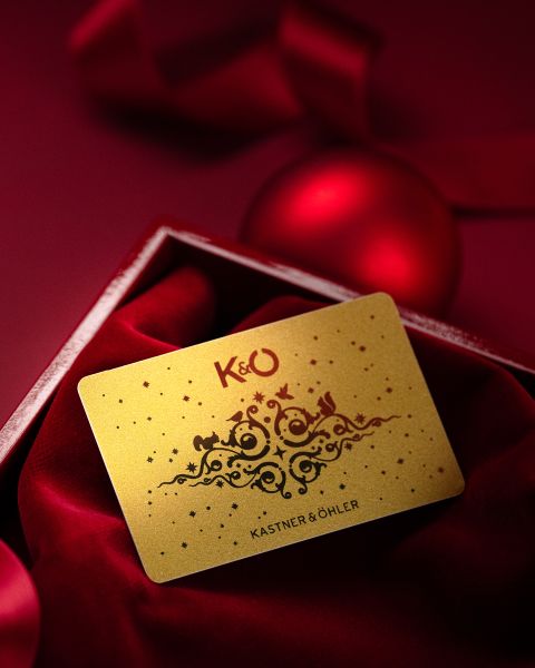Eine goldene, mit festlichen Mustern verzierte Kastner & Öhler-Geschenkkarte liegt auf rotem Samt in einer offenen Schachtel, mit roten Ornamenten im Hintergrund.
