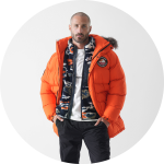 Ein Mann steht mit dem Gesicht nach vorne und trägt eine leuchtend orangefarbene Pufferjacke mit einer pelzbesetzten Kapuze, einen gemusterten Fleece, eine schwarze Hose und ein weißes T-Shirt vor einem schlichten weißen Hintergrund.