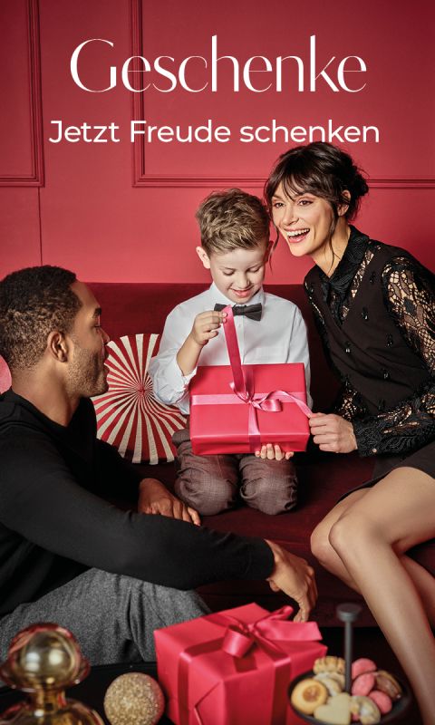 Eine lächelnde dreiköpfige Familie sitzt auf einem roten Sofa. Ein kleiner Junge öffnet eine rote Geschenkschachtel, während seine Eltern fröhlich zusehen. Der Hintergrund und die Dekoration sind festlich, darüber steht der Text "Geschenke Jetzt Freude schenken".