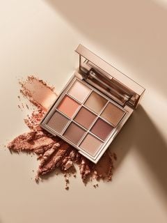 Eine quadratische Make-up-Palette mit neun Lidschatten in neutralen Tönen liegt offen auf einer beigen Fläche, auf der einige der pudrigen Farbtöne verstreut und zerdrückt sind. Im Deckel der Palette ist ein Spiegel zu sehen.