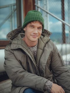 Ein Mann, der eine grüne Strickmütze, einen grauen Pullover und eine olivgrüne Winterjacke trägt, sitzt im Freien auf Holzstufen und lächelt sanft in die Kamera. Im Hintergrund sind ein Metallzaun und ein Glasgebäude zu sehen.