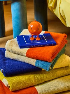 Ein Stapel bunter Handtücher, jedes in kräftigen Blöcken in Orange, Blau, Gelb und Beige, mit einem blauen Buch und einer kleinen orangefarbenen, runden Figur obenauf. Die Szene befindet sich in einem Innenraum auf einem Holzfußboden.
