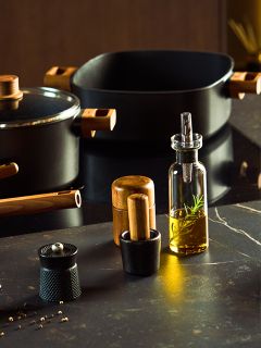 Eine moderne Küchenarbeitsplatte zeigt schwarzes Kochgeschirr mit Holzgriffen, eine Glasflasche mit Rosmarinöl, eine Pfeffermühle, eine Salzmühle und einen kleinen Mörser mit Stößel, alles auf einer dunklen Fläche.
