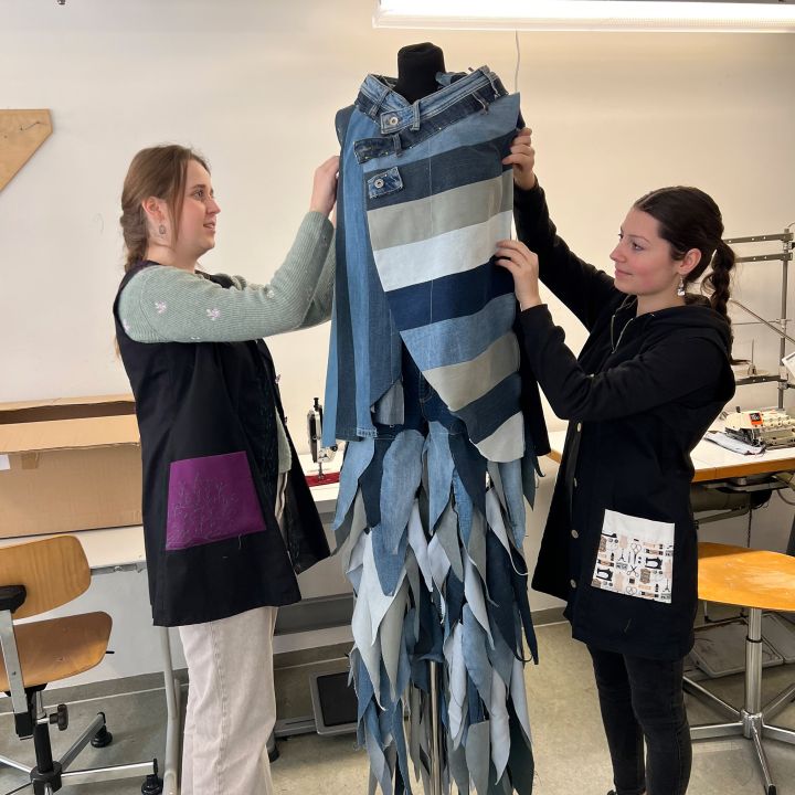 Zwei Frauen passen ein Jeanskleidungsstück mit gestreiften und gezackten Mustern auf einer Kleiderform in einer Werkstatt an, umgeben von Stühlen und Nähmaschinen.