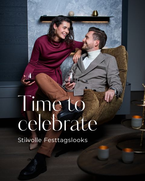 Ein lächelndes Paar in eleganten Kleidern genießt gemeinsam Wein in einem Haus. Die Frau in einem burgunderroten Kleid sitzt auf einem Schrank, und der Mann in einem hellen Blazer sitzt auf einem braunen Stuhl. Der Text lautet: "Time to celebrate. Stilvolle Festtagslooks.".