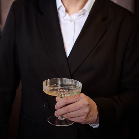 Eine Person in schwarzem Anzug und weißem Hemd hält ein leeres, strukturiertes Glasgefäß, wobei nur ihr Oberkörper und ihre Hand vor einem dunklen Hintergrund zu sehen sind.