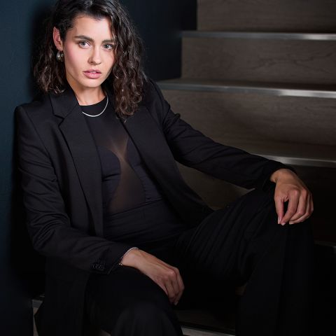 Eine Frau mit gewelltem braunem Haar, die einen schwarzen Anzug und ein durchsichtiges schwarzes Oberteil trägt, sitzt auf einer Holztreppe vor einer dunklen Wand und schaut selbstbewusst in die Kamera.