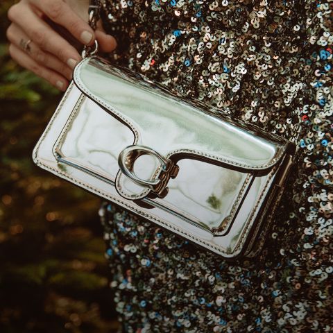 Eine Person hält eine metallisch glänzende, silberne Handtasche mit Schnallenverschluss in der Hand und trägt ein paillettenbesetztes Kleid mit schwarzen und silbernen Pailletten.