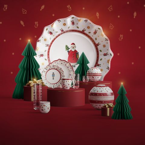 Ein festliches Geschirrset mit Tellern, Tassen und Gläsern mit weihnachtlichen Motiven, umgeben von dekorativen grünen Papierbäumen, Geschenkboxen und Ornamenten, alles vor einem roten Hintergrund.