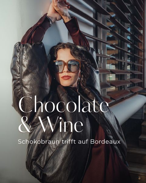 Eine elegante Frau mit Sonnenbrille und dunkler Pufferjacke posiert im Freien vor einem modernen Gebäude. Der Text lautet: "Schokolade & Wein. Schokobraun trifft auf Bordeaux.