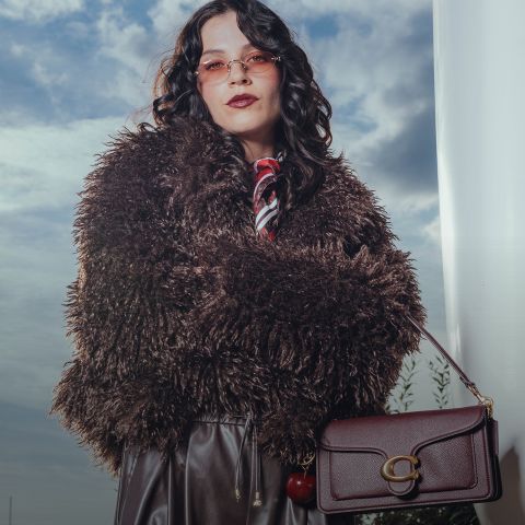 Eine Person mit gewelltem dunklem Haar trägt eine rosa getönte Brille, einen wuscheligen braunen Mantel, eine dunkle Lederhose und einen roten Schal. Sie hält eine weinrote Handtasche und steht im Freien vor einem bewölkten Himmel.