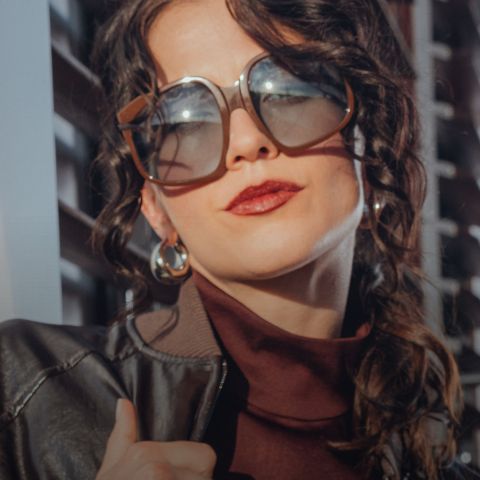 Eine Frau mit gewelltem dunklem Haar, einer übergroßen Sonnenbrille, Ohrringen, einem kastanienbraunen Rollkragenpullover und einer dunklen Lederjacke posiert selbstbewusst im Freien.