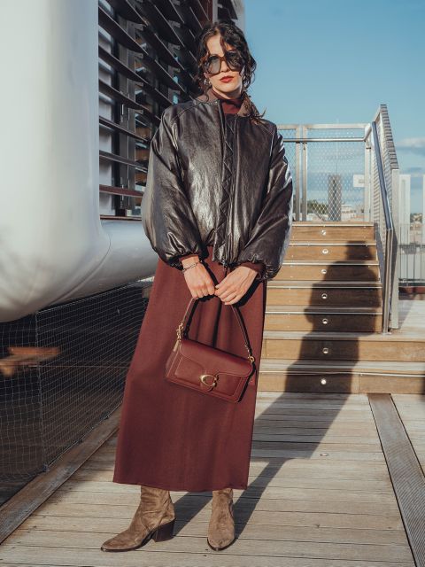 Eine Frau steht im Freien auf einer Holztreppe, trägt ein langes weinrotes Kleid, eine glänzende schwarze Jacke, hellbraune Wildlederstiefel, eine Sonnenbrille und eine passende weinrote Handtasche. Der Himmel ist klar und blau.