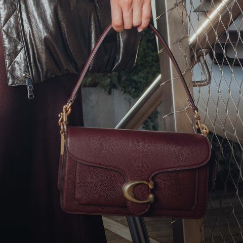 Eine Person in einer dunklen, glänzenden Jacke hält eine weinrote Lederhandtasche mit einem goldenen "C"-Verschluss und einem kurzen Henkel in der Hand.