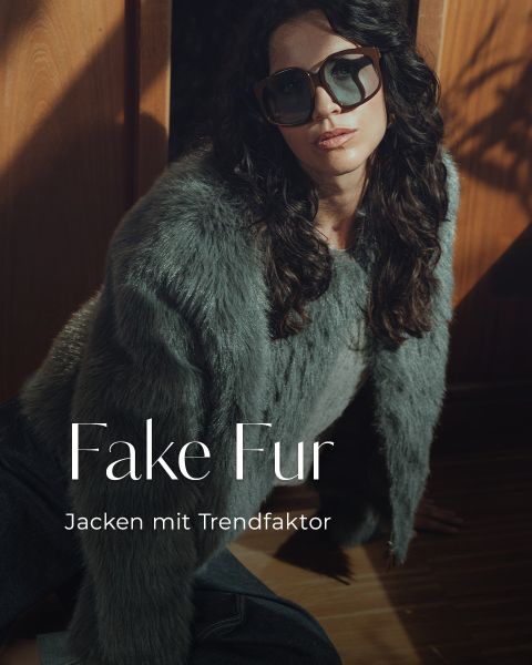 Eine Frau mit gewelltem dunklem Haar und großer Sonnenbrille trägt eine graue Kunstpelzjacke. Sie posiert in einem Innenraum bei dramatischer Beleuchtung. Der überlagerte Text lautet: "Fake Fur. Jacken mit Trendfaktor.