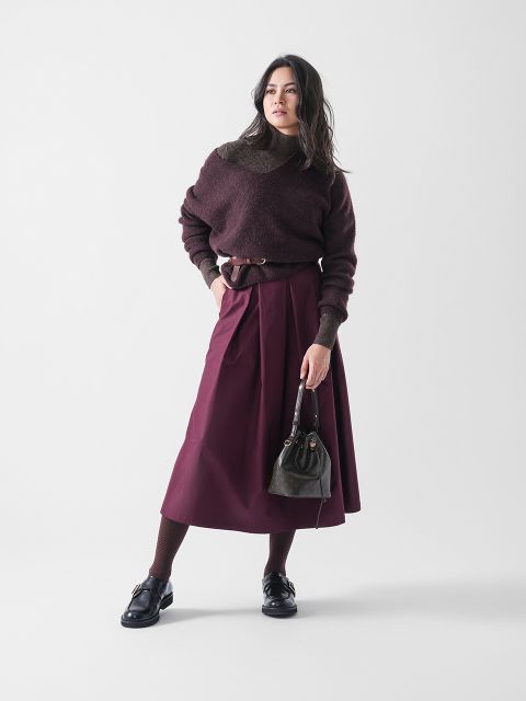 Eine Frau steht vor einem schlichten Hintergrund und trägt einen dunklen kastanienbraunen Pullover, einen passenden Midirock mit Gürtel, eine gemusterte Strumpfhose, schwarze Schuhe und eine kleine dunkle Handtasche. Sie hat langes dunkles Haar und einen selbstbewussten Gesichtsausdruck.