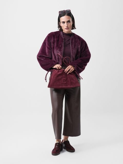 Eine Person steht vor einem schlichten weißen Hintergrund und trägt eine weinrote Kunstpelzjacke, eine braune Hose mit weitem Bein und weinrote Turnschuhe. Sie hält eine passende burgunderfarbene Handtasche und trägt eine Sonnenbrille auf dem Kopf.