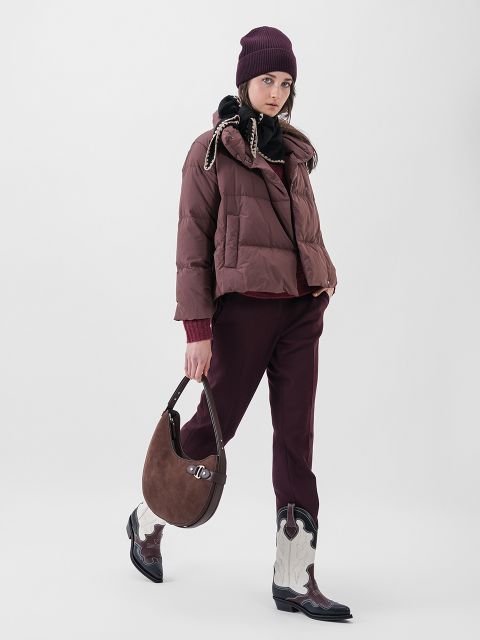 Eine Person, die eine kastanienbraune Pufferjacke, eine passende Mütze, einen dunklen Schal, eine weinrote Hose und schwarz-weiße Cowboystiefel trägt und eine braune Handtasche in der Hand hält, steht vor einem schlichten hellen Hintergrund.