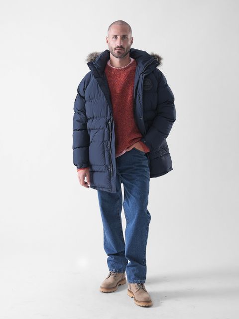 Ein Mann steht vor einem schlichten weißen Hintergrund und trägt eine marineblaue Pufferjacke mit einer pelzgefütterten Kapuze, einen roten Pullover, blaue Jeans und hellbraune Schuhe.