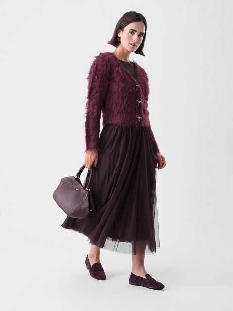 Eine Frau in einem lila Pullover und Rock, die eine Handtasche hält.