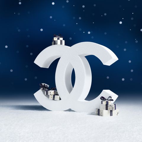 Weißes Chanel-Logo mit kleinen, eingepackten Geschenkschachteln vor einer verschneiten Fläche und einem dunkelblauen, verschneiten Hintergrund.