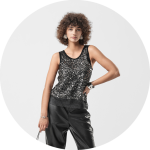 Eine Frau mit lockigem Haar posiert in einem ärmellosen schwarzen Paillettenoberteil und einer schwarzen Lederhose, in der Hand eine kleine Handtasche, vor einem schlichten hellen Hintergrund.