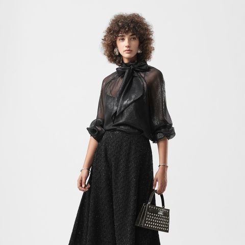 Frau mit lockigem Haar in schwarzer, durchsichtiger Bluse mit Schleife und langem, strukturiertem schwarzem Rock, die eine mit Nieten besetzte Handtasche in der Hand hält, steht vor einem schlichten Hintergrund.
