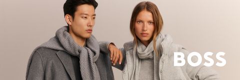 Zwei Models in grauen, modischen Winteroutfits mit Pullovern und Schals posieren vor einem neutralen Hintergrund. Das Wort "BOSS" erscheint in fetten weißen Buchstaben.