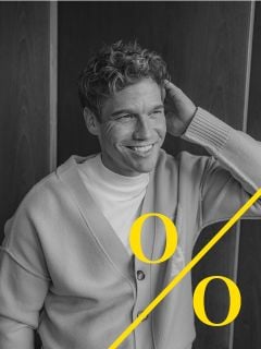 Ein Mann mit lockigem Haar, der eine helle Strickjacke und ein weißes Hemd trägt, sitzt lächelnd da und hält eine Hand hinter den Kopf. Das Schwarz-Weiß-Bild zeigt zwei große gelbe "o "s und eine diagonale gelbe Linie am unteren Rand.