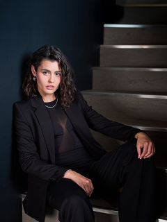 Eine Frau in einem schwarzen Anzug sitzt auf einer Holztreppe vor einer dunklen Wand und blickt selbstbewusst in die Kamera, ihr Haar ist zu lockeren Wellen gestylt.