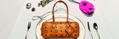 Eine braune Designer-Handtasche von MCM steht auf einem weißen Teller, umgeben von Silberbesteck, einem Maßband, einem metallischen Ornament und einem rosafarbenen, wuscheligen Accessoire, das einen gedeckten Esstisch imitiert.