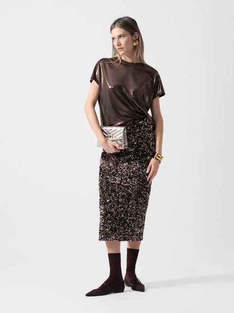 Frau trägt ein glänzendes braunes Oberteil, einen glitzernden Paillettenrock, kastanienbraune Socken, schwarze Schuhe und hält eine Metallclutch vor einem weißen Hintergrund.