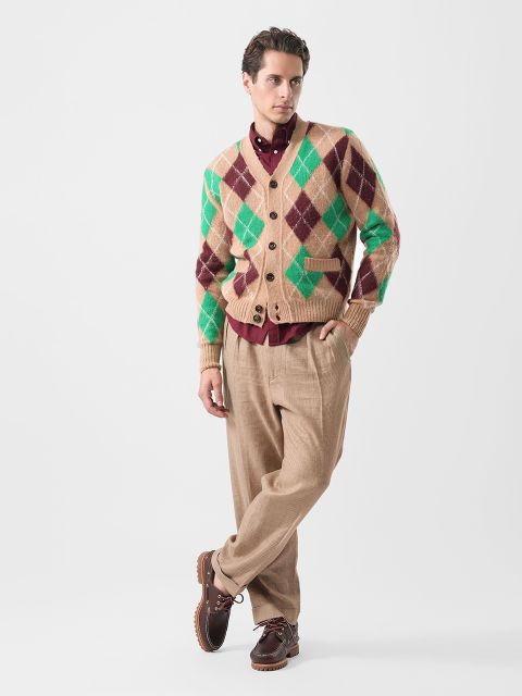 Ein Mann posiert in einer grün-braunen Strickjacke, einem burgunderroten Hemd, einer beigen Hose und braunen Schuhen vor einem schlichten weißen Hintergrund.