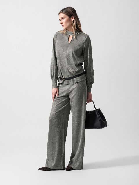Eine Frau trägt eine silbern schimmernde Bluse und eine dazu passende Hose mit weitem Bein, hält eine schwarze Handtasche in der Hand und blickt vor einem schlichten Hintergrund zur Seite.