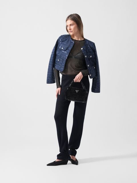 Eine Frau in einer strukturierten blauen Jacke, einer schwarzen Hose und schwarzen Flats hält eine schwarze Handtasche vor einem schlichten weißen Hintergrund.