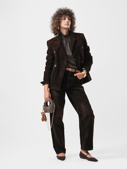 Eine Person mit lockigem Haar trägt einen dunkelbraunen Samtanzug, eine Metallbluse und braune flache Schuhe und hält eine kleine Tasche mit Anhängern vor einem schlichten Hintergrund.