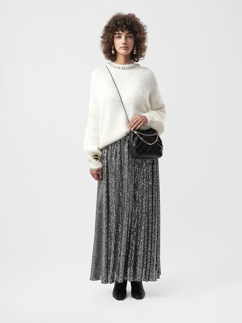 Frau mit lockigem Haar trägt einen weißen Pullover, einen gemusterten Maxirock, schwarze Stiefel, goldene Ohrringe und eine schwarze Umhängetasche vor einem schlichten Hintergrund.