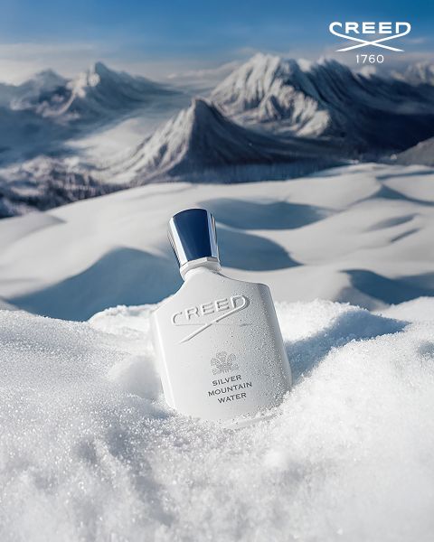 Ein weißer Creed Silver Mountain Water Parfümflakon liegt im Schnee mit verschneiten Berggipfeln im Hintergrund unter einem blauen Himmel.