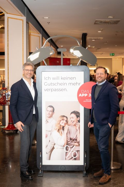 Zwei Männer in Anzügen stehen neben einem großen Schild in einem Geschäft und werben für eine App mit einem Foto von drei lächelnden Frauen und einem Mann auf dem Plakat.