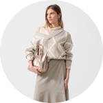 Frau in einem hellbeigen Pullover und Rock, mit einer Metallhandtasche in der Hand, mit offenem Haar und Statement-Ohrringen, vor einem schlichten Hintergrund.