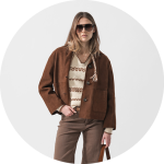 Frau mit brauner Jacke, beigefarbenem gemustertem Pullover, brauner Hose und Sonnenbrille, stehend vor einem schlichten hellen Hintergrund.