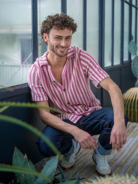 Ein Mann mit lockigem Haar in einem rosa gestreiften Hemd und Jeans hockt in einem Haus in der Nähe von Pflanzen und lächelt in die Kamera.