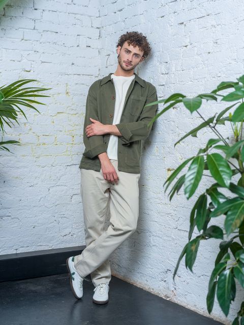 Ein Mann mit lockigem Haar in grüner Jacke, weißem Hemd und beiger Hose lehnt an einer weißen Backsteinmauer, umgeben von Grünpflanzen.