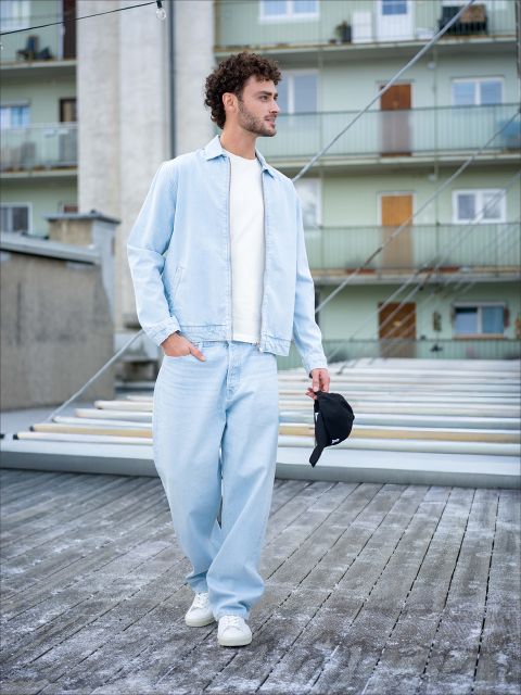 Ein Mann in hellblauen Jeans, passender Jacke und weißen Turnschuhen geht im Freien spazieren und hält eine schwarze Mütze in der Hand, im Hintergrund sind Wohnhäuser zu sehen.