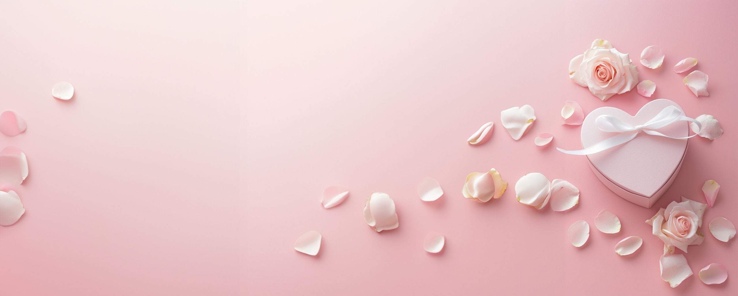 Herzförmiger Geschenkkarton mit weißem Band, Rosen und gestreuten Rosenblättern auf zartrosa Hintergrund.