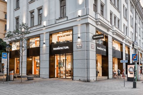 Ein modernes Eckgeschäft mit großen Fenstern und schwarzen Schildern mit der Aufschrift "MANUFACTUM". Draußen auf dem Bürgersteig laufen Menschen vorbei.