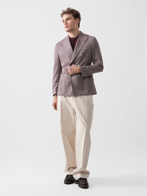 Ein Mann, der einen malvenfarbenen Zweireiher, ein dunkles Hemd und eine weite cremefarbene Hose trägt, steht vor einem schlichten weißen Hintergrund.