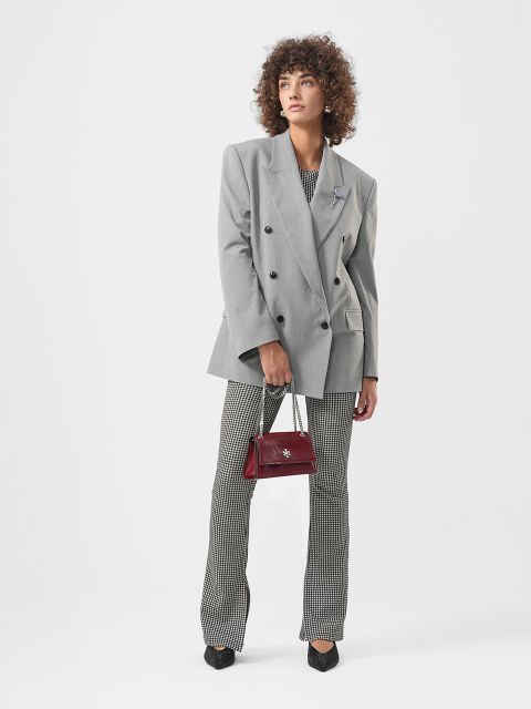 Eine Frau in einem grauen übergroßen Blazer, einer gemusterten Hose und schwarzen Schuhen hält eine kleine weinrote Handtasche und steht vor einem schlichten weißen Hintergrund.
