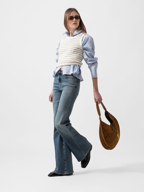 Frau mit Sonnenbrille, blau gestreiftem Hemd, weißer Strickweste, Schlagjeans und schwarzen Schuhen, hält eine braune Umhängetasche und steht selbstbewusst da.